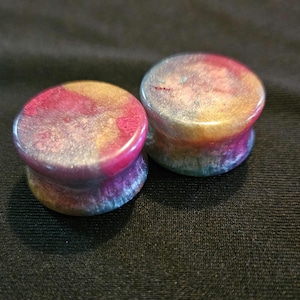 May include: Two colorful ear gauges with a marbled effect. The plugs feature a mix of pink, gold, blue, and purple hues. The jewelry is cylindrical with a flared design, and the surface has a glossy finish. The gauges are set against a dark background.