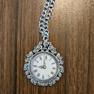 Puede incluir: Un collar de plata con un colgante de reloj. La esfera del reloj es blanca con números romanos negros y una aguja plateada. El colgante está rodeado por un marco decorativo de plata con detalles florales.