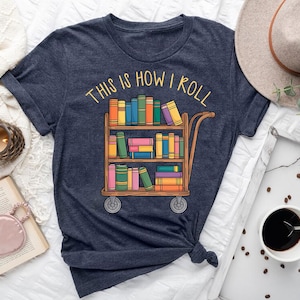 May include: Navy blue t-shirt with the text "THIS IS HOW I ROLL" above a graphic of a wooden cart filled with colorful books. The cart has two wheels and is filled with books of various colors.