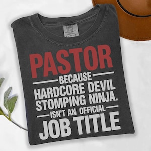 May include: Dark gray t-shirt with the word "PASTOR" in red block letters. Below, white text reads "BECAUSE HARDCORE DEVIL STOMPING NINJA. ISN'T AN OFFICIAL JOB TITLE."