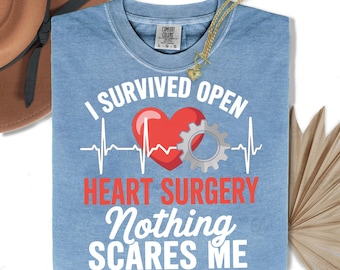 May include: A light blue t-shirt with the text "I SURVIVED OPEN HEART SURGERY Nothing SCARES ME" in white and red. The design includes a red heart, a silver gear, and a white EKG line. A gold necklace with a heart pendant is draped over the shirt.