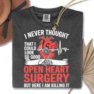 May include: Dark gray t-shirt with the text "I NEVER THOUGHT THAT I COULD LOOK SO GOOD After OPEN HEART SURGERY BUT HERE I AM KILLING IT." The shirt features a red anatomical heart graphic with a medical device and a white EKG line.