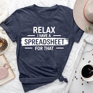 May include: A navy blue t-shirt with the white text "RELAX I HAVE A SPREADSHEET FOR THAT". The shirt is knotted at the bottom. A beige hat, coffee cup, and other items are in the background.