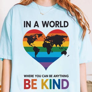 May include: Light blue t-shirt with the text "IN A WORLD WHERE YOU CAN BE ANYTHING BE KIND." The shirt features a rainbow-colored heart with a world map silhouette.