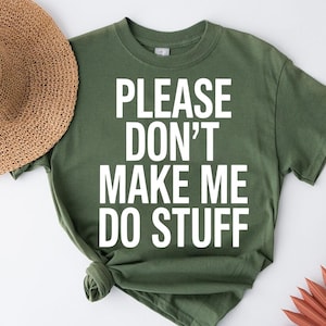 May include: Olive green t-shirt with the text "PLEASE DON'T MAKE ME DO STUFF" in white block letters. A straw sun hat is partially visible on the left. The shirt is knotted at the bottom. A decorative fan is in the bottom right corner.