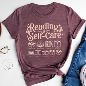 a purple t-shirt with the text "Reading is Self-Care" printed on it, along with various illustrations of books and reading-related objects. The t-shirt is placed on a white surface, and there are some other items, such as a pair of jeans, a pair of glasses, and a pair of sneakers, visible in the background.