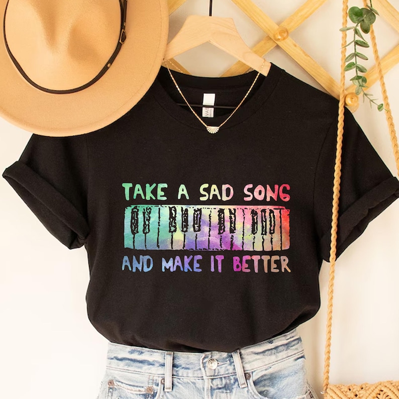 A black t-shirt with a colorful graphic design and text printed on it, hanging on a wooden hanger.