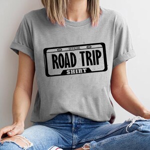 A person wearing a gray t-shirt with a graphic design of a license plate and the text "ROAD TRIP SHIRT" printed on it.