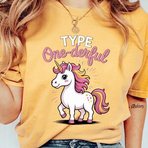 May include: Mustard yellow t-shirt with a cartoon unicorn design. The unicorn has a pink and yellow mane and tail, and a golden horn. The text "TYPE One-derful" is above the unicorn. The unicorn is walking.