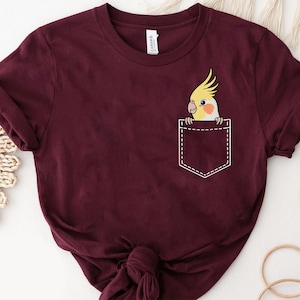 Op de afbeelding: Bordeauxrood T-shirt met een zakontwerp met een cartoon kaketoe die eruit piekt. De kaketoe heeft gele kuifveren, een perzikwangkleur en een wit gezicht. De zak is wit omlijnd en het shirt heeft een ronde hals.