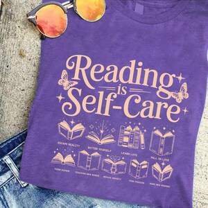 A purple t-shirt with the text "Reading is Self-Care" printed on it, along with illustrations of books and butterflies. The t-shirt is placed on a surface with a pair of sunglasses and a pair of jeans visible in the background.