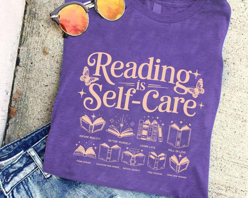 A purple t-shirt with the text "Reading is Self-Care" printed on it, along with illustrations of books and butterflies. The t-shirt is placed on a surface with a pair of sunglasses and a pair of jeans visible in the background.