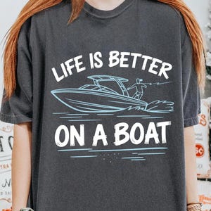May include: Dark gray t-shirt with the text "LIFE IS BETTER ON A BOAT" in white and a light blue illustration of a speedboat with a person waterskiing. The shirt is a crew neck.
