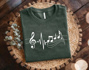 May include: Dark green t-shirt featuring a white musical note design. The design includes a treble clef, a heartbeat-like line, and musical notes on a staff. The shirt is displayed on a woven basket.