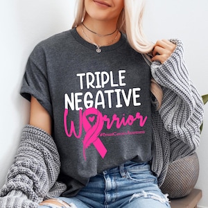 A woman wearing a gray t-shirt with the text "Triple Negative Warrior" and a pink ribbon graphic, along with a necklace and a gray sweater.