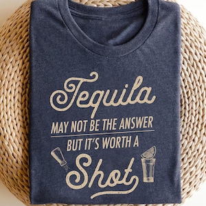 Op de afbeelding: Marineblauw t-shirt met de tekst "Tequila may not be the answer but it's worth a shot" in een decoratief lettertype. Het ontwerp bevat illustraties van een zoutstrooier en een tequila shot met een limoenpartje.