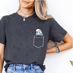 以下が含まれることがあります： 漫画のセキセイインコのポケットデザインが施された、濃いグレーのTシャツ。セキセイインコは青、白、黄色です。シャツは柔らかい素材でできており、半袖です。