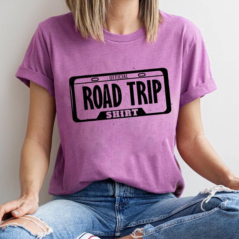 A person wearing a purple t-shirt with a graphic design of a license plate and the text "ROAD TRIP SHIRT" printed on it.