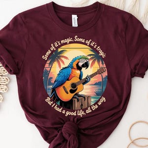 May include: Burgundy t-shirt featuring a colorful parrot playing a guitar against a tropical sunset backdrop. The design includes the text "Some of it's magic, Some of it's tragic, But I had a good life, all the way."