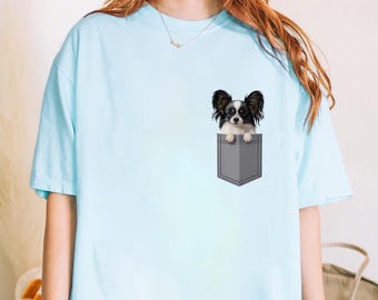 Könnte beinhalten: Hellblaues T-Shirt mit einem Aufdruck eines Papillon-Hundes, der aus einer grauen Tasche schaut. Der Hund hat schwarz-weißes Fell und ist auf der Brust zentriert. Das T-Shirt hat kurze Ärmel und einen Rundhalsausschnitt.