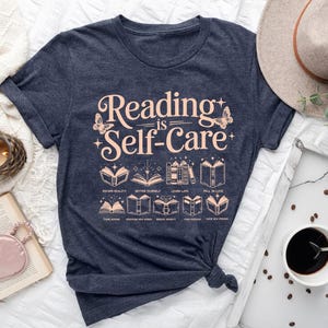 a blue t-shirt with the text "Reading is Self-Care" printed on it, along with illustrations of books and other related items. The t-shirt is placed on a bed, surrounded by various objects such as a hat, a cup of coffee, and a book.