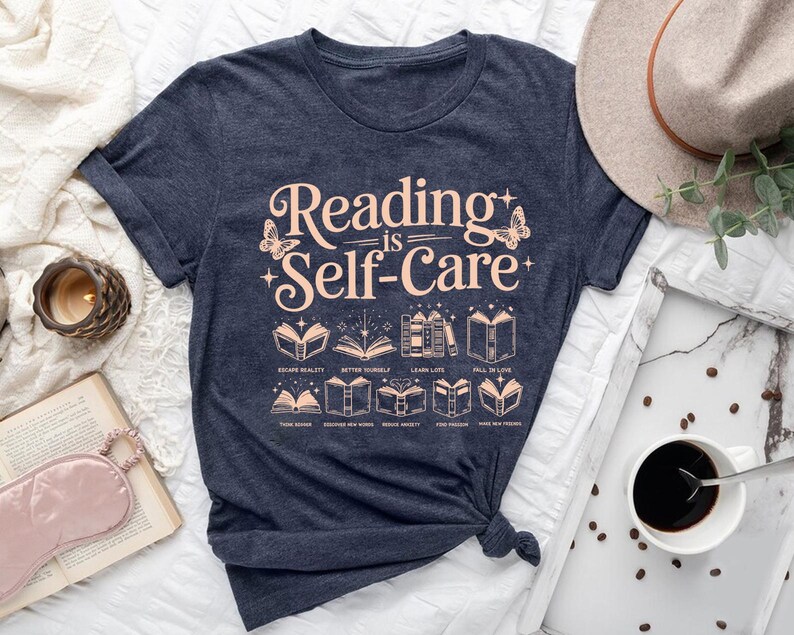 a blue t-shirt with the text "Reading is Self-Care" printed on it, along with illustrations of books and other related items. The t-shirt is placed on a bed, surrounded by various objects such as a hat, a cup of coffee, and a book.