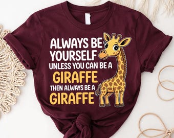 May include: A maroon t-shirt with the text "ALWAYS BE YOURSELF UNLESS YOU CAN BE A GIRAFFE THEN ALWAYS BE A GIRAFFE" in white and yellow. A cartoon giraffe is on the right side of the shirt.