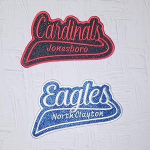 May include: Two embroidered patches with glittery finishes. The top patch is black with red text reading Cardinals and Jonesboro. The bottom patch is blue with white text reading Eagles and North Clayton. Both feature a swoosh design element.