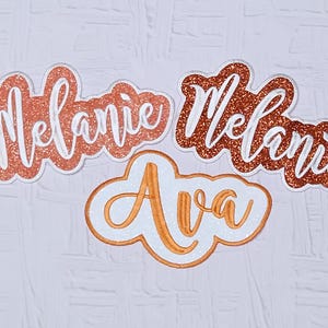 May include: Three glittery name cutouts. Two read "Melanie" in a copper-colored script with a white border. The third cutout says "Ava" in a similar style. All are on a white background.