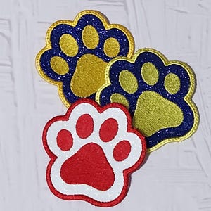 May include: Three embroidered paw print patches. Two are blue and gold with glitter accents, and one is red with a white border and glitter accents. These patches are likely for pet lovers or crafting projects.