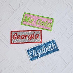 May include: Three rectangular embroidered patches with glittery backgrounds. One pink patch reads "Mz. Cola" in green, another silver patch reads "Georgia" in red, and a blue patch reads "Elizabeth" in white.