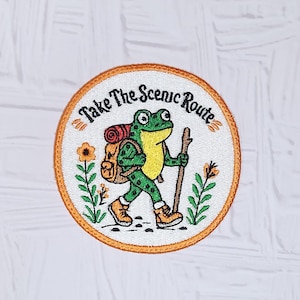 May include: A circular embroidered patch with a white background and an orange border. It features a green frog wearing a brown backpack and hiking boots, holding a wooden walking stick. The text "Take The Scenic Route" is arched above.