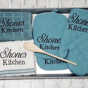May include: A set of kitchen towels and oven mitts with the text "Shone's Kitchen" embroidered on them. The set includes two towels, one white and one blue, and two oven mitts, both blue. A wooden spoon is also included.