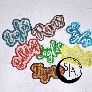 May include: A collection of seven iron-on patches with different school mascot names. The patches are in various colors, including red, blue, green, yellow, and brown. The names on the patches include "Eagles", "Rams", "Bulldogs", "Tigers", and "Wildcats".