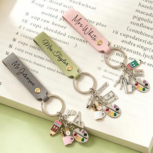 May include: Three personalised leather keychains in grey, sage green, and light pink. Each features a metal ring with charms shaped like a pencil, a cup of pens, a paint palette, and a ruler. The straps are engraved with Mr. Johnson, Mrs. Taylor, and Mrs. White.