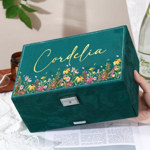 May include: A teal jewelry box with the name "Cordelia" in gold script. The box features a floral border with yellow, pink, and white flowers. It has a silver clasp and a rectangular shape.
