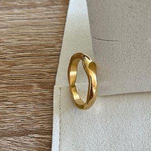 Könnte beinhalten: Ein goldfarbener Ring mit einem einzigartigen, fließenden Design. Der Ring wird auf einer weißen, strukturierten Oberfläche präsentiert, mit dem Markennamen "STILECHT COLOGNE" sichtbar. Die Form des Rings ist fließend und organisch.