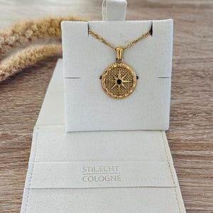 Pode incluir: Um colar de ouro com um pingente de b&uacute;ssola que apresenta uma estrela preta no centro. O colar &eacute; exibido em uma caixa de veludo branco com o texto "Stil.Echt Cologne" na parte inferior.