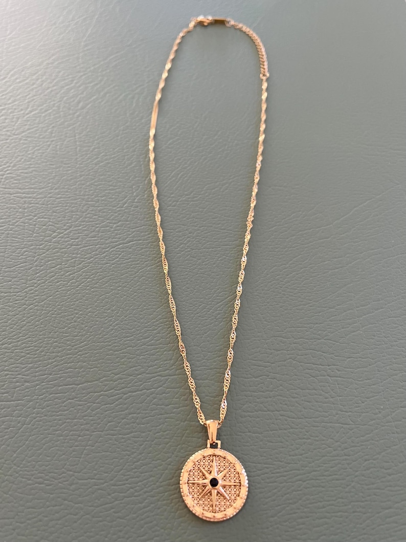 Pode incluir: Um colar de corrente de ouro com um pingente de b&uacute;ssola. A b&uacute;ssola &eacute; redonda e tem um centro preto com uma estrela dourada.