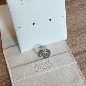 May include: A pair of silver hoop earrings in a white velvet jewelry box with the text "Stil.Echt Cologne" printed on the bottom.