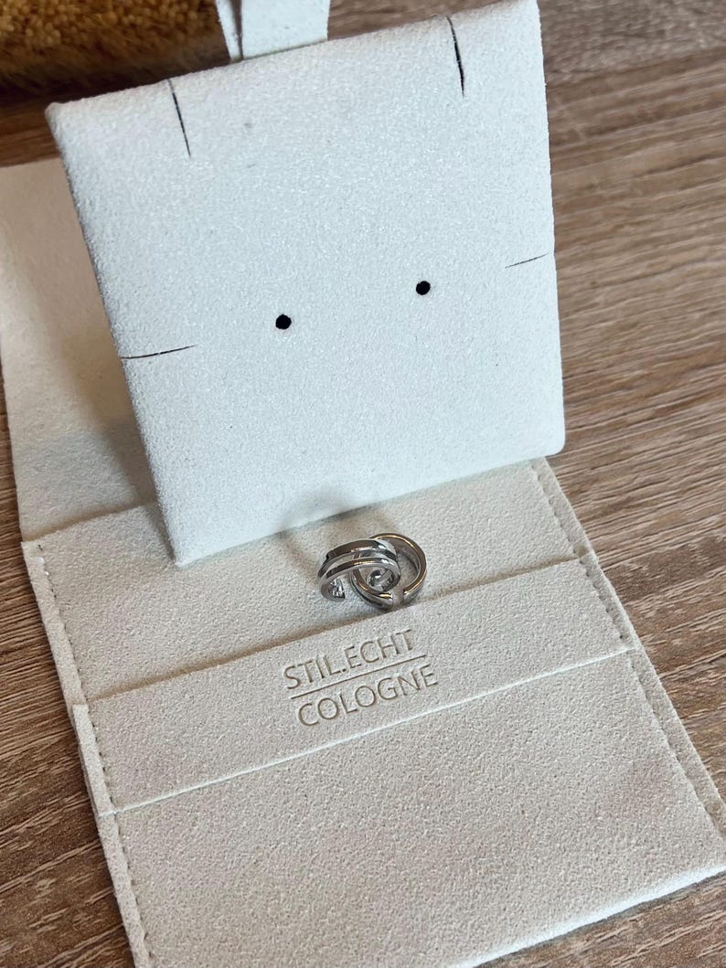 May include: A pair of silver hoop earrings in a white velvet jewelry box with the text "Stil.Echt Cologne" printed on the bottom.