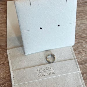 May include: A white felt jewelry box with a silver ring inside. The box has the text "STIL.ECHT COLOGNE" printed on the inside.