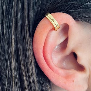 May include: Gold ear cuff with a simple, minimalist design. The cuff is made of two thin, flat pieces of metal that are joined together at the top. The cuff is worn on the upper part of the ear, just above the cartilage.