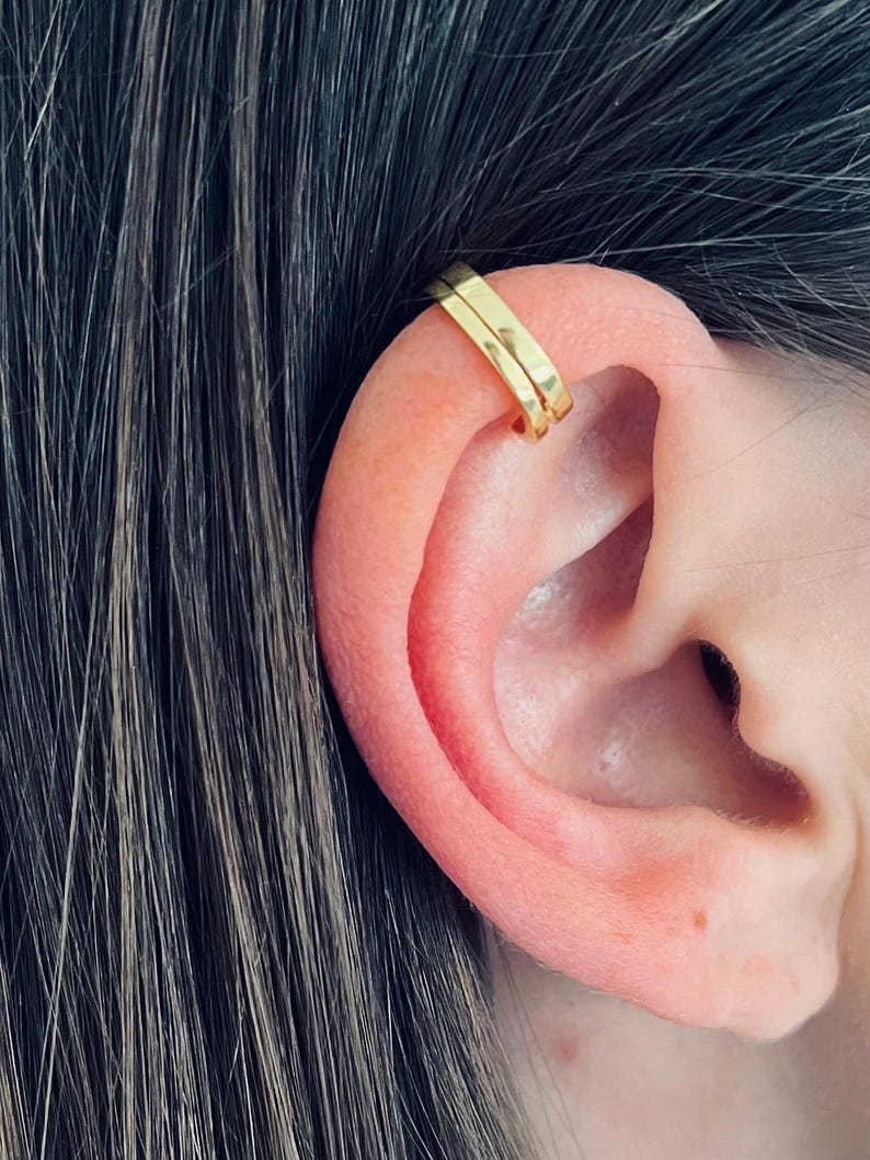 May include: Gold ear cuff with a simple, minimalist design. The cuff is made of two thin, flat pieces of metal that are joined together at the top. The cuff is worn on the upper part of the ear, just above the cartilage.