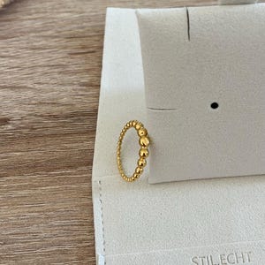 Könnte beinhalten: Ein goldfarbener Ring mit Perlenmuster, der auf einer hellgrauen Präsentationskarte liegt. Der Ring hat eine Reihe kleiner, zusammengefasster Perlen. Auf der Präsentationskarte steht der Text "STILECHT COLOGNE".