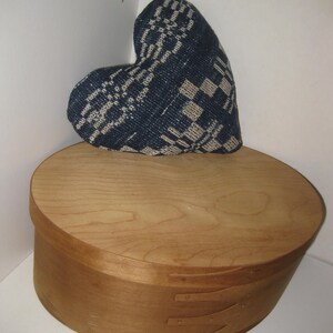 May include: A heart-shaped pillow with a blue and white woven pattern rests on a light brown wooden oval box. The box has a lid and is constructed with visible dovetail joints, showcasing a rustic aesthetic.