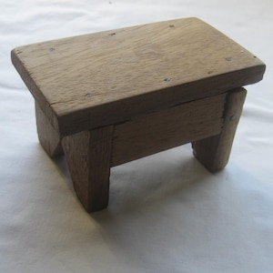 May include: A small, handcrafted wooden stool with a rectangular seat and four sturdy legs. The stool is made of light brown wood, showing visible grain and natural imperfections. It appears to be a rustic, handmade item, perfect for decorative or functional use.