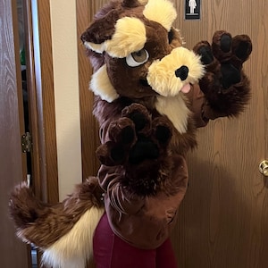 Może przedstawiać: Brązowy i beżowy kostium fursuit, z głową, ciałem i długim ogonem, połączony z czerwonymi spodniami. Kostium ma duże łapy i przyjazny wyraz twarzy, zaprojektowany tak, aby przypominał postać z kreskówki.