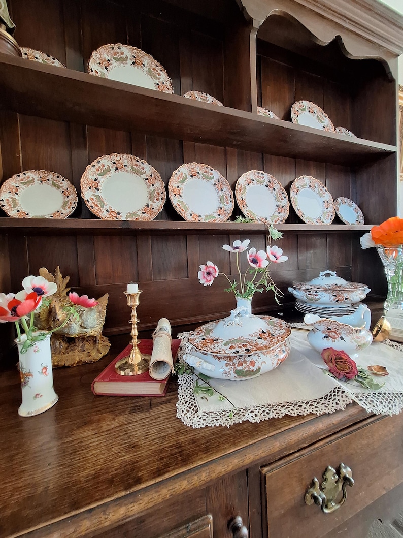 Victorian-style dresser display featuring antique Crown Derby Imari china, covered vegetable tureens, gravy boat, decorative plates, trailing ivy, garden flowers and old books creating a classic heritage interior vignette.