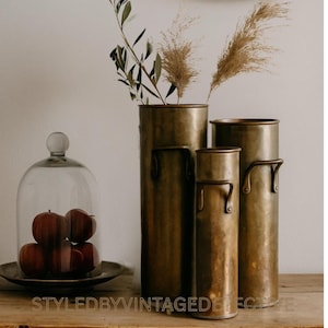 May include: A still life arrangement featuring four bronze cylindrical vases of varying heights, with handles. The vases hold dried grasses and leafy branches. A glass cloche with red apples sits on a wooden surface. The text "STYLEDBYVINTAGEDECOR" is visible.
